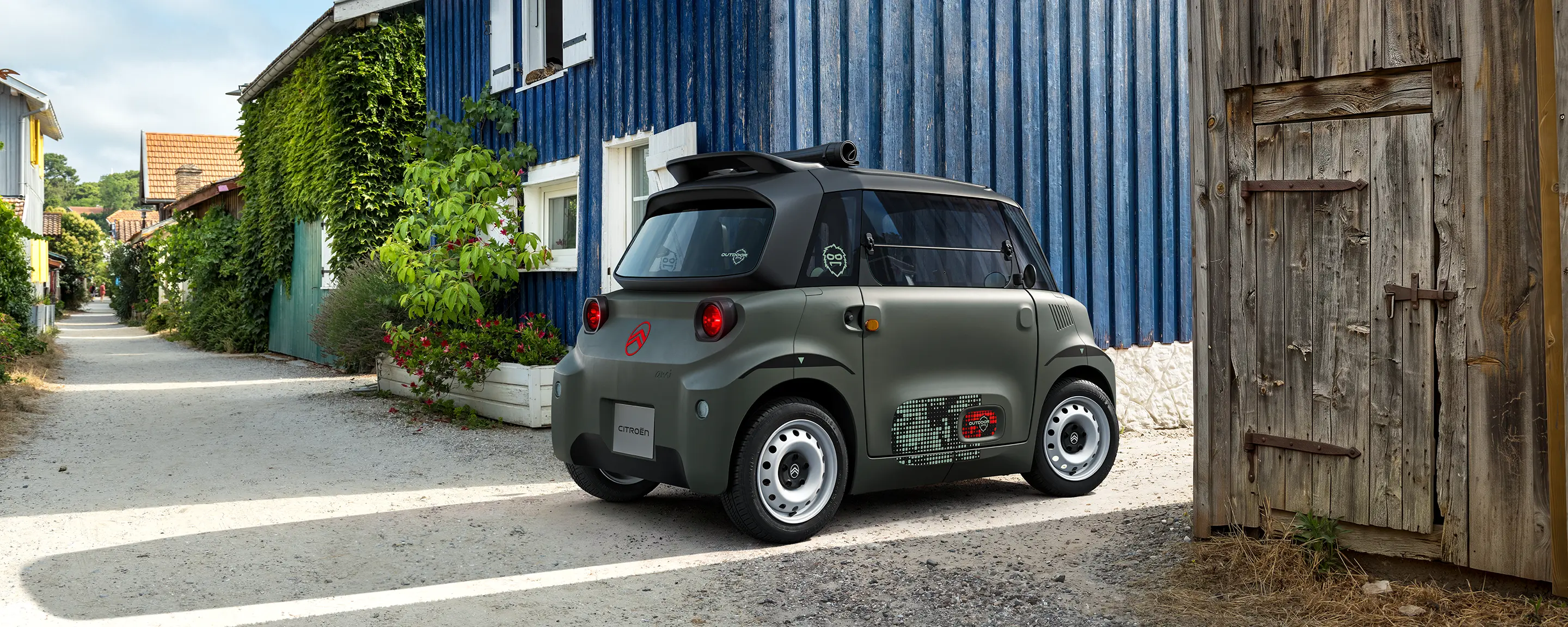 Vista anteriore a tre quarti della Citroën Ami Outdoor con fari LED, una microcar elettrica compatta in un borgo costiero